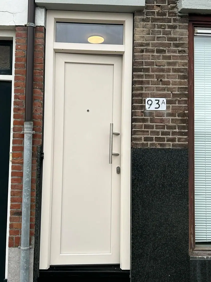 Crèmekleurige kunststof voordeur met bovenlicht en rvs handgreep in een bakstenen gevel.